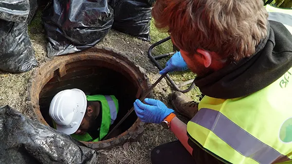 Two drainage professionals working on a blocked drain, one wearing a white hard hat and green safety vest inside a manhole, while the other holds a tool above, surrounded by black bags.