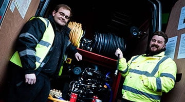 Two drainage specialists in high-visibility jackets standing beside drainage equipment in a service van, showcasing expertise in emergency drain services for stadiums and event spaces.
