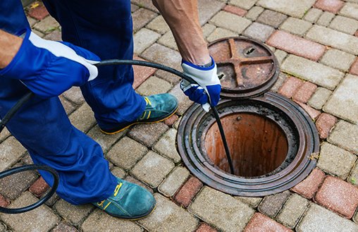 Person in blue gloves using a drain cleaning tool on a manhole, illustrating common causes of blocked drains and the need for professional drainage services.