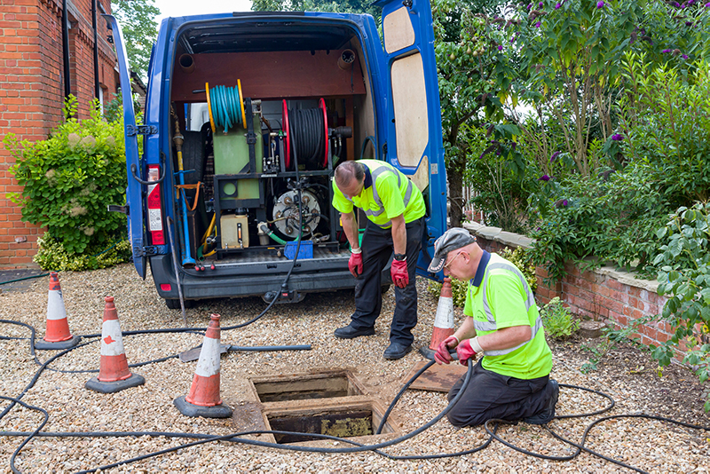 Two drainage technicians in high-visibility clothing working near an open drain, with a van equipped for drain unblocking in the background, showcasing tools and equipment for drainage services.