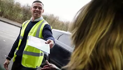 Smiling man in a high-visibility vest handing a document to a woman, representing Environ Drainage Services' customer-focused approach to emergency drain services for stadiums and events.