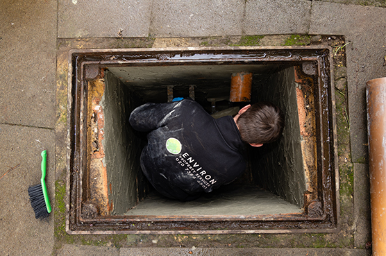 Technician from Environ Drainage Services inspecting a drainage system in a manhole, with tools like a brush nearby, highlighting commercial drainage solutions.